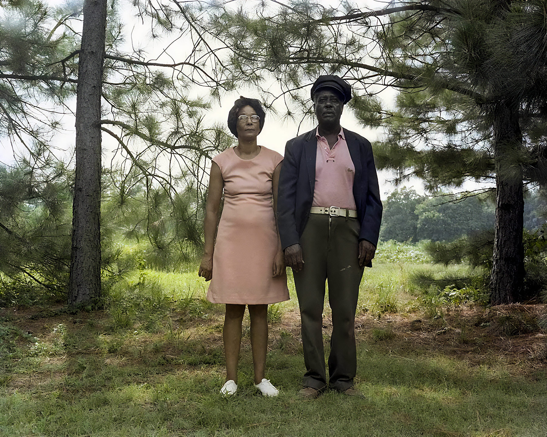 A color portrait of Jessie Mae and Jasper Staples, a Black couple standing for a portrait in formal attire in front of tall pine trees.