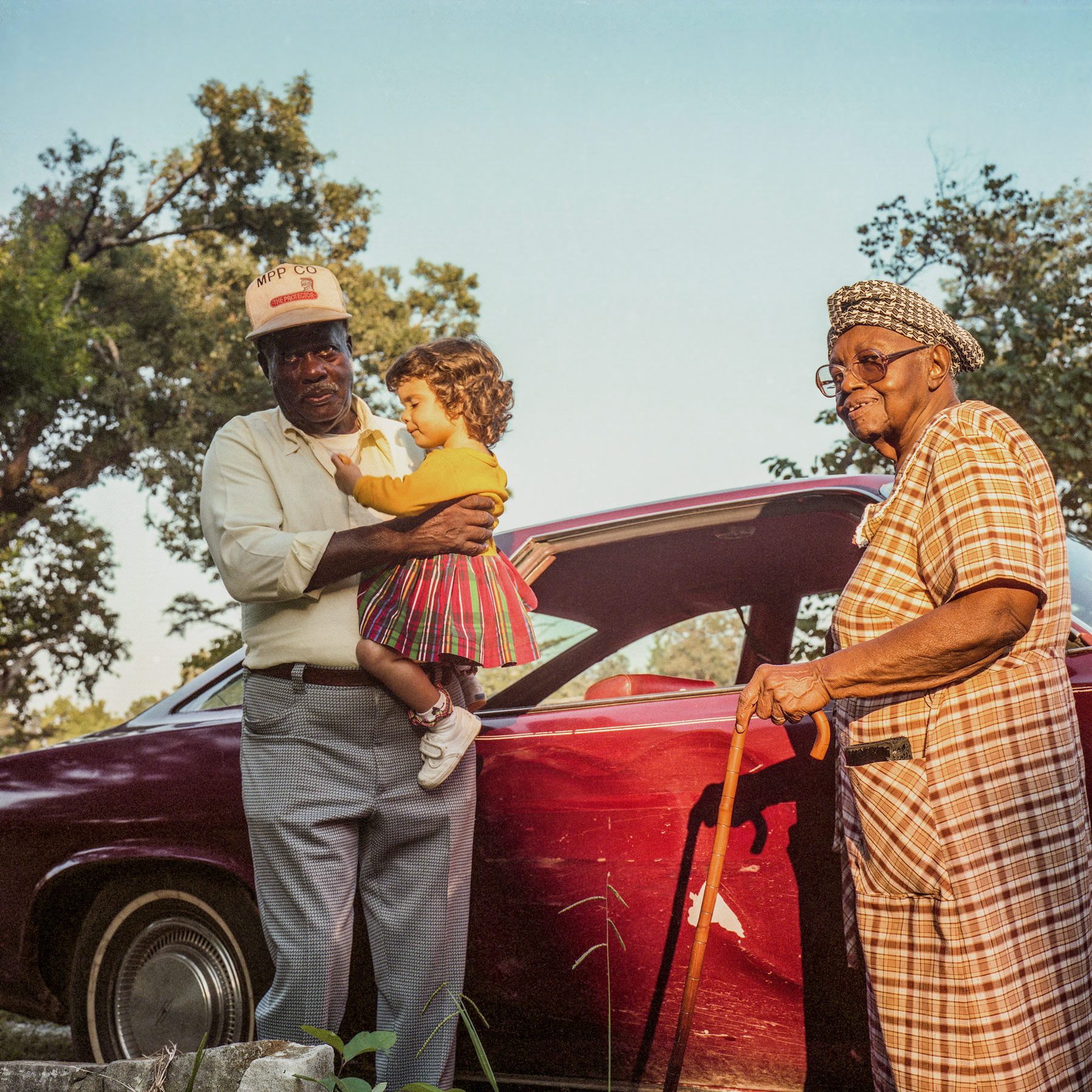 A square color portrait of Jasper Staples, an older Black man with a hat and button down shirt, holding a young white girl. To the right of them is Lucille Fleming, an older Black woman with a cane. They both stand in front of a red car.