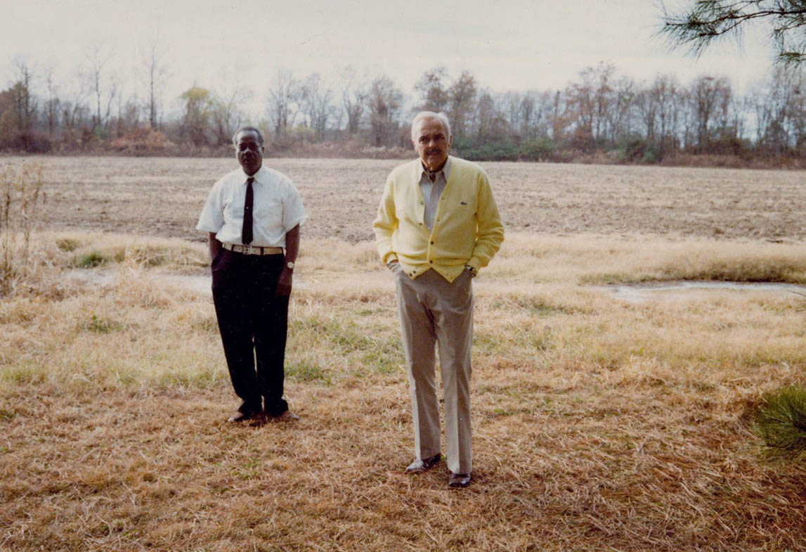 A color portait of Jasper Staples, an older Black man wearing a white button down and tie, and Adyn Schuyler, an older white man wearing a button down and yellow cardigan. Jasper stands to the left and behind Adyn. Both are in front of a fallow field.