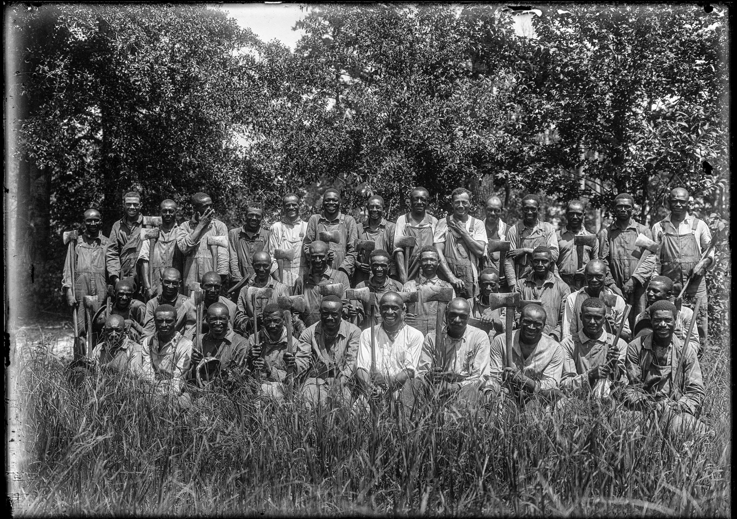 A black and white portrait of 36 Black men in three rows wearing workwear and holding axes, posing for a portrait in a field.