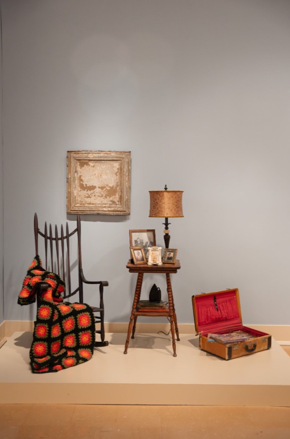 Christina McField (born 1992), The Table Remembers and Echoes in the Tin, 2025; Installation view; Found tin ceiling tile, antique rocking chair, small wooden table, antique suitcase filled with books, picture frames, reproductions of family photographs, small table lamp, antique pressing iron, found crocheted blanket