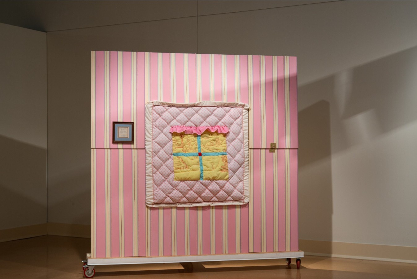 Rylee Brabham (born 2001), a breeze or a breath, 2025; Installation view; Plywood, two-by-fours, steel, casters, paint, trim, old bedsheet, yellow napkins, red thread, clamps, oscillating fan, heart-shaped tooth fairy box, windchime, faucet handle, pink plastic, empty testosterone bottle, clown hands, needlepoint, lightswitch plate, steel, pink flock, plastic baby