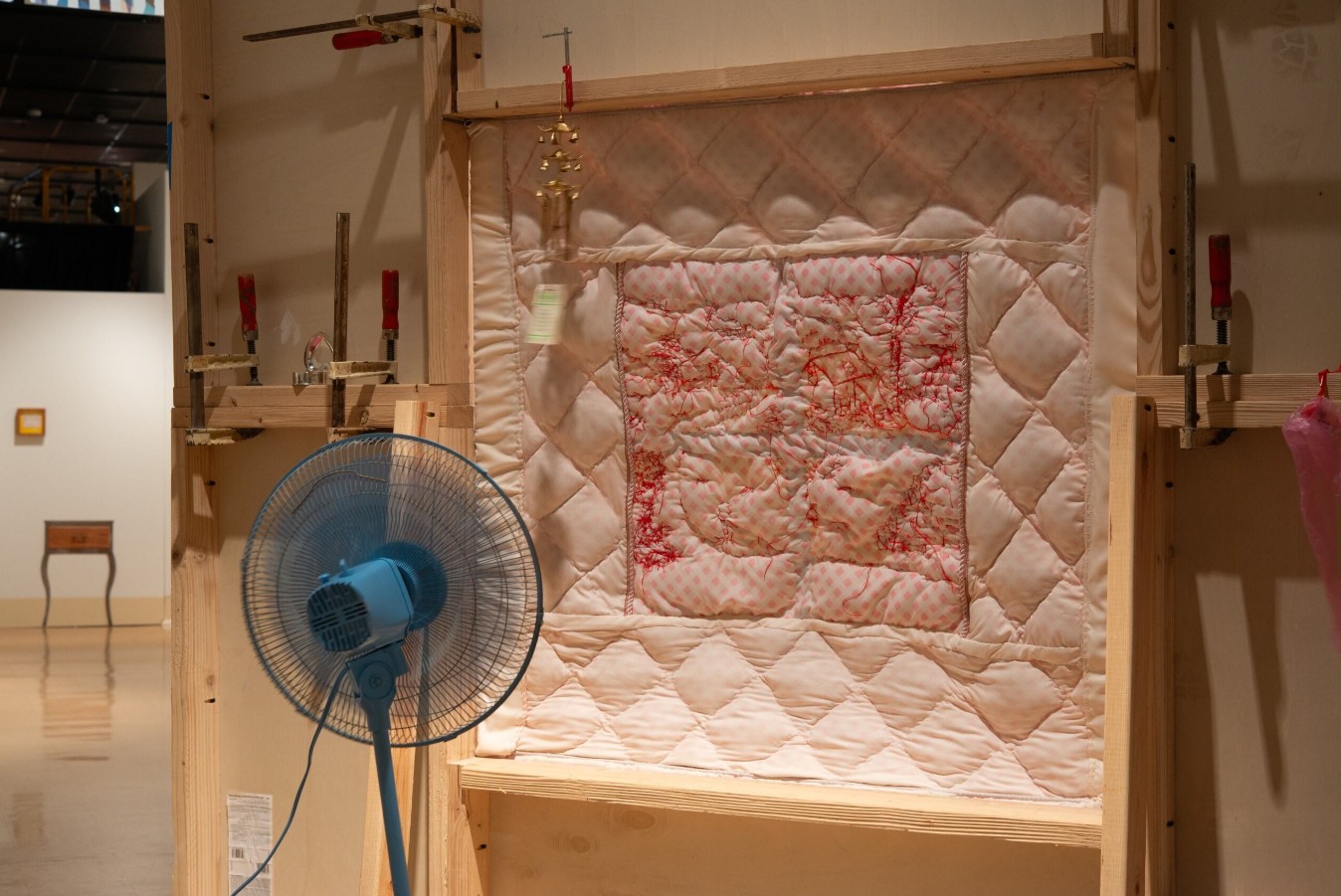 Rylee Brabham (born 2001), a breeze or a breath, 2025. Installation view. Plywood, two-by-fours, steel, casters, paint, trim, old bedsheet, yellow napkins, red thread, clamps, oscillating fan, heart-shaped tooth fairy box, windchime, faucet handle, pink plastic, empty testosterone bottle, clown hands, needlepoint, lightswitch plate, steel, pink flock, plastic baby